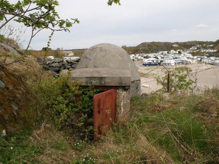 Liten betongstruktur med rusten metallport, omgitt av vegetasjon, med hvite campingvogner parkert i bakgrunnen på et sandete område.