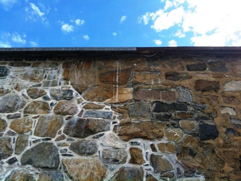 Steinmur på Akershusfestning med ulike størrelser og farger på steinene, noen uregelmessig formet, med synlig mørtel mellom steinene, under en klar blå himmel med spredte hvite skyer.