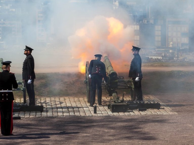 Uniformerte militærpersonell som opererer en kanon i ferd med å avfyres, med stor flamme og røyk synlig ved munningen, på et asfaltert område med gress og bygninger i bakgrunnen.