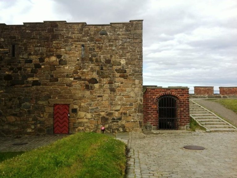 Steinbygning med rød dør og lite vindu, ved siden av en buet murstruktur med svart metallport, brosteinssti og trapper som fører til et forhøyet område med gress og en annen steinmur i bakgrunnen, under en overskyet himmel.