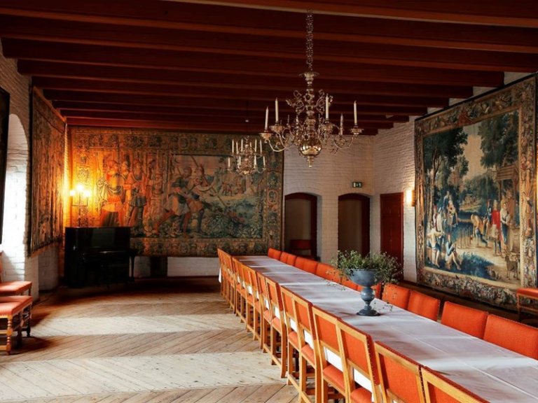 Vakker sal med lang bord dekket med hvit duk, omgitt av oransje stoler, med trebjelker i taket, to utsmykkede lysekroner, store veggtepper med historiske motiver, og et piano i hjørnet.