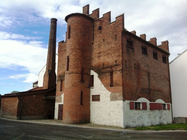 Gamle murbygning med sylindrisk tårn og krenelerte brystvern, med en blanding av røde murstein og hvitmalte seksjoner, smale vinduer og høy skorstein i bakgrunnen.