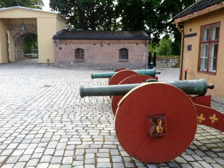 Brosteinsbelagt gårdsplass med to historiske kanoner montert på røde trevogner, foran en liten murbygning med buede vinduer og en større struktur med en buegang, omgitt av trær.