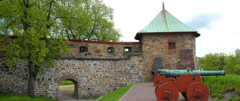 Historiske Munks tårn med grønnaktig tak, buet inngang i muren, og to gamle kanoner på trevogner, omgitt av grønt gress og trær.