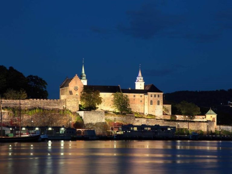 Akershus festning i Oslo, Norge, opplyst om natten med refleksjon i vannet i forgrunnen, som fremhever den historiske arkitekturen og steinmurene.