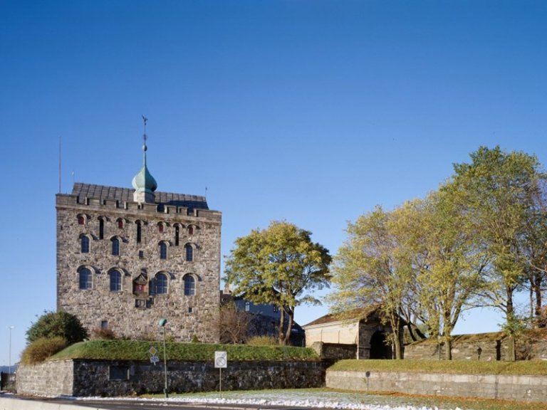Historisk steinbygning med grønn kuppel og buede vinduer, omgitt av trær og klar blå himmel.