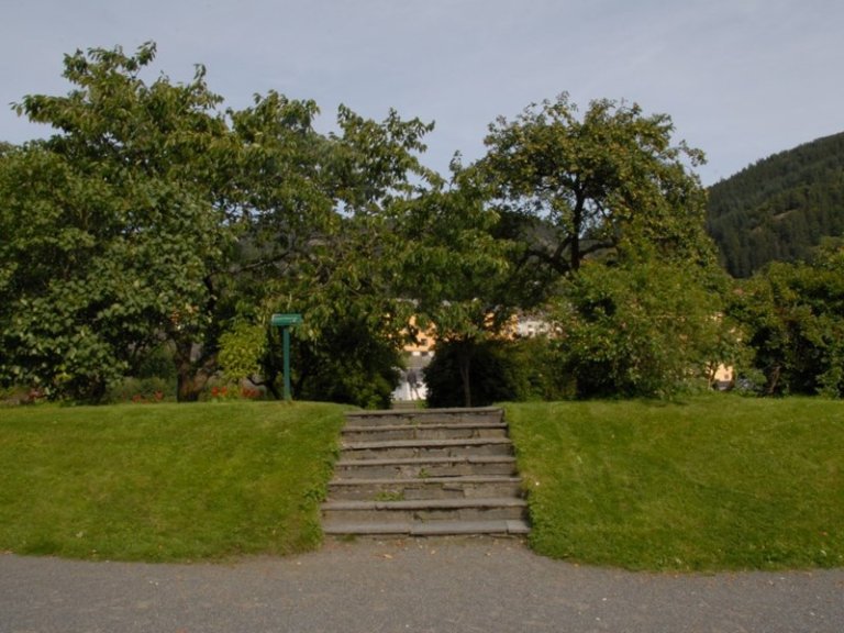Liten steintrapp som fører opp til et gressområde med trær og busker, med flere trær og bygninger delvis skjult av løvverk i bakgrunnen, og et fjell synlig i det fjerne på høyre side under en klar himmel.