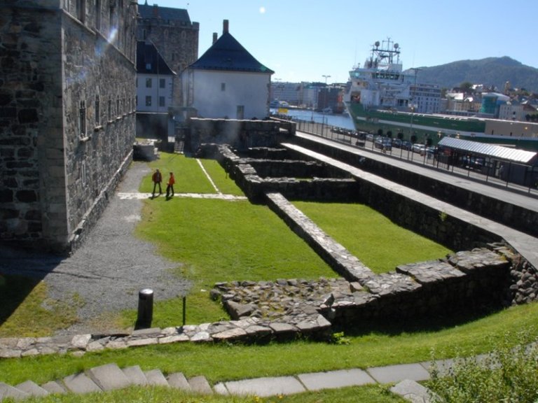 Utendørsområde med steinruiner og gressplen ved siden av en stor steinbygning, med to personer som går på stien. I bakgrunnen er det en havn med et stort skip og bygninger langs vannkanten på en solrik dag.