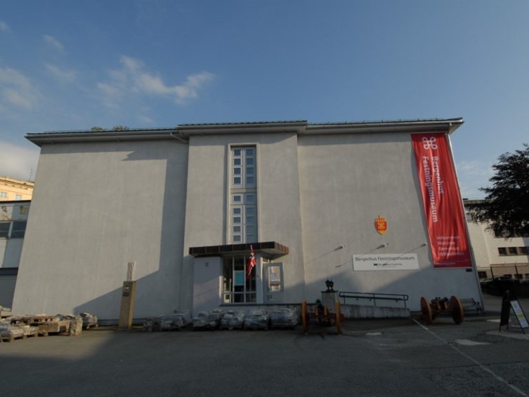 Moderne bygning med flatt tak og lys grå farge, med inngang med glassdører og vinduer, norsk flagg ved inngangen, og et stort rødt banner som leser 'Bergenhus Festningsmuseum'.