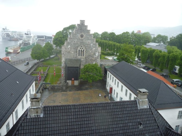 Historisk steinbygning med gavltak og buede vinduer, omgitt av moderne bygninger, trær og grøntområder, med en havn i bakgrunnen på en overskyet dag.