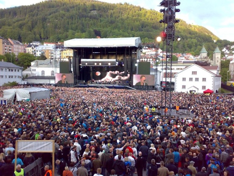 Stor utendørskonsert med et massivt publikum foran en scene, utstyrt med store skjermer og artister, med bygninger og en skogkledd åsside i bakgrunnen.