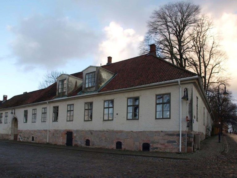 Stor, gammel bygning med hvite vegger og rødt tak, flere vinduer og to kvistvinduer, steinbunn og buet døråpning, omgitt av brosteinsbelagt område og høye trær uten blader i bakgrunnen.