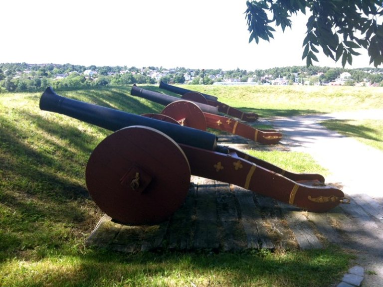 Rekke med fire historiske kanoner på en gresskledd plass, sannsynligvis en del av et gammelt fort eller militærinstallasjon, med trær og bygninger i bakgrunnen.