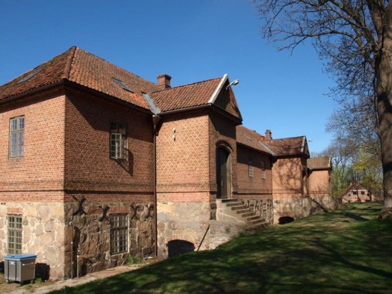 Stor murbygning med takstein, flere vinduer og steinfundament, med inngang og trapp, omgitt av gress og trær.