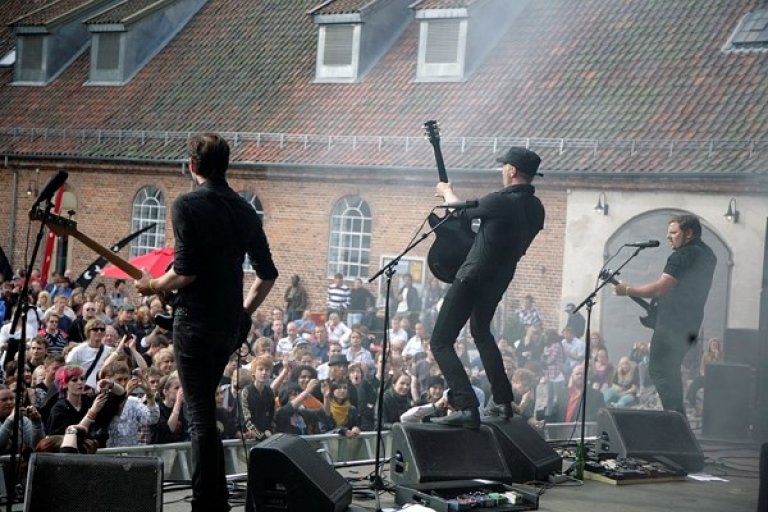 Live musikkopptreden med tre musikere på scenen, hvor den ene musikeren spiller elektrisk gitar og beveger seg energisk, med et stort publikum foran scenen og en bygning med flere vinduer og takstein i bakgrunnen.