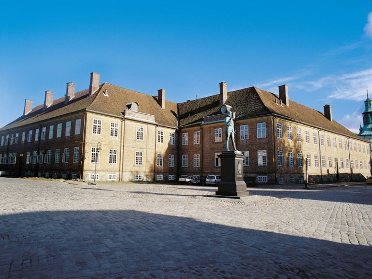 Stor, historisk bygning med statue foran, flere vinduer og skorsteiner, brosteinsbelagt område og noen parkerte biler, under en klar blå himmel.