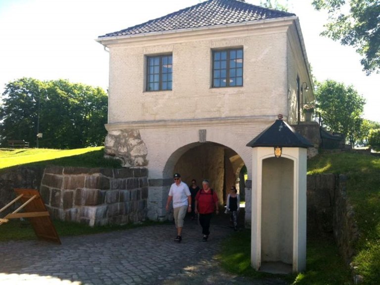 Gruppe mennesker som går gjennom en buegang av en historisk bygning med hvite vegger, blå innrammede vinduer og takstein, med en liten vaktbod til høyre og steinvegger og grøntområder rundt.