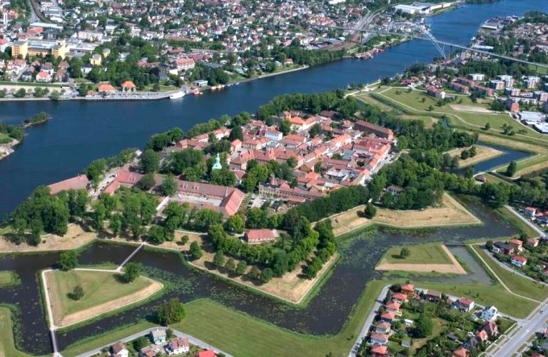 Luftfoto av en historisk festning med stjerneformet layout, omgitt av vann og grøntområder, med boliger, veier og vannveier i nærheten.