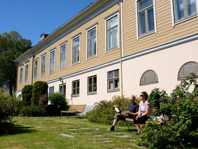Toetasjes bygning med beige kledning og hvite detaljer, med gressplen, busker og trær foran, og to personer som sitter på en benk under en klar blå himmel
