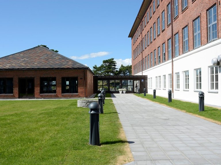 Uteområde med to bygninger, en enetasjes murbygning til venstre og en fleretasjes mur- og hvitmalt bygning til høyre, med en asfaltert gangvei mellom dem