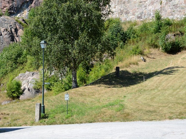 Uteområde med gressplen, tre, busker og en lyktestolpe ved siden av et skilt, med en steinete høyde og steinmur i bakgrunnen.