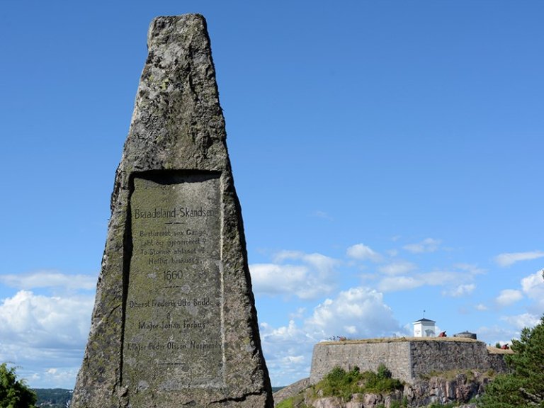 Steinmonument med inskripsjon på norsk, med en festning på en høyde i bakgrunnen under en blå himmel med noen skyer