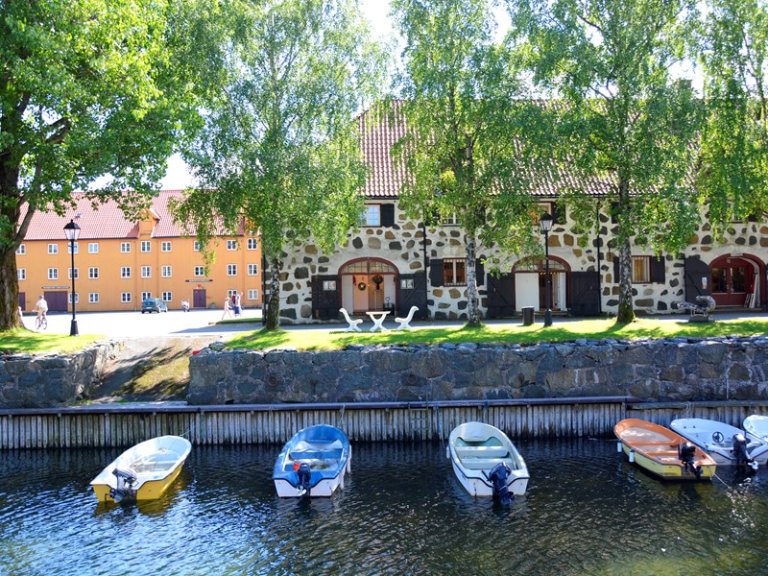 Vannfront med små båter ved kai, steinbygning med buede dører og rødt tak, og gul bygning med vinduer i bakgrunnen.