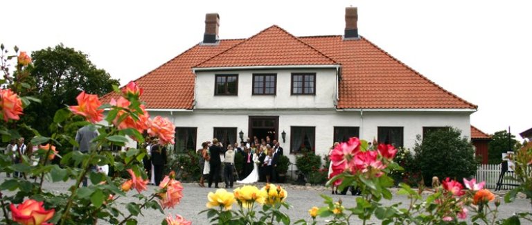 Bryllupsfest foran Kommandantboligen en hvit bygning med rødt tak og vakre blomster i forgrunnen.