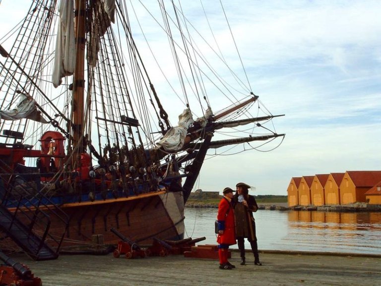 Stor, gammeldags seilskute ved kai med to personer i historiske klær og gule bygninger med røde tak i bakgrunnen.