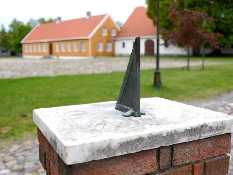 Solur montert på mursteinsokkel i utendørs omgivelser, med to bygninger med røde tak og gule og hvite vegger i bakgrunnen, omgitt av gress og brosteinsstier.