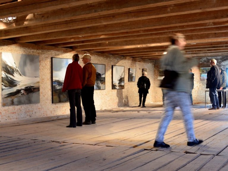 Kunstgalleri med flere personer som observerer malerier på veggene, med rustikk interiør, trebjelker i taket og tregulv, og varm belysning som skaper en koselig atmosfære.