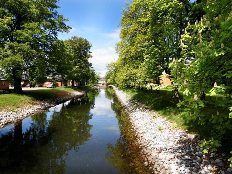 Fredelig kanal omgitt av frodige grønne trær og steinbelagte bredder, med bygninger i bakgrunnen og blå himmel reflektert i vannet.