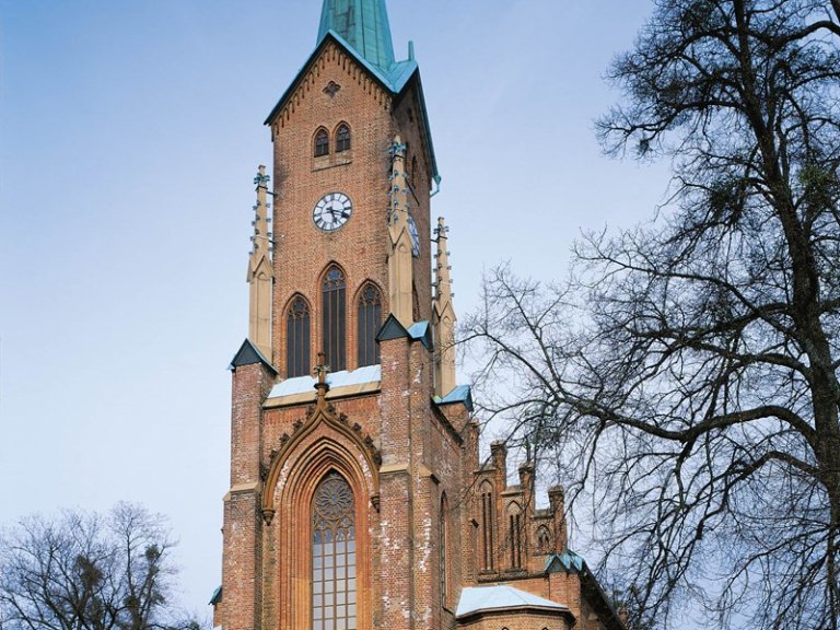 Høy murkirketårn med klokke på forsiden, med gotiske arkitektoniske elementer som spisse buer og dekorative spir, omgitt av nakne trær og klar blå himmel.