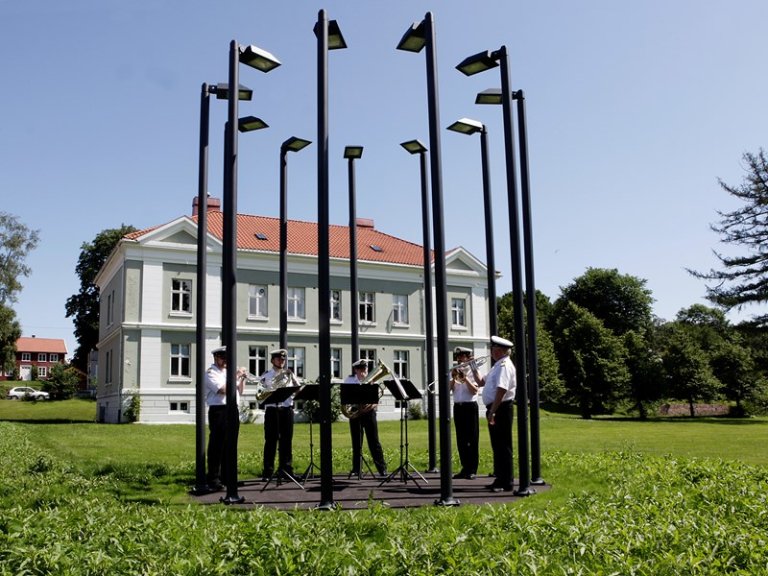 Et brassband spiller musikk utendørs, omgitt av høye lyktestolper, foran en stor hvit bygning med rødt tak på en solrik dag.