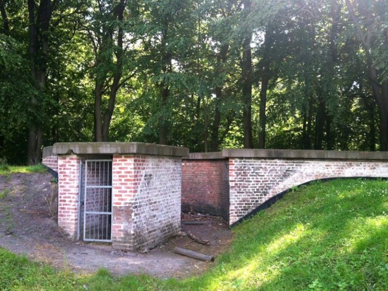 Liten murstruktur med metallgitterdør, delvis innebygd i bakken, omgitt av jordhaug og skogområde.