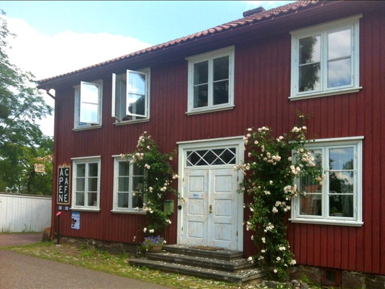 To-etasjes rød trebygning med hvitmalte vinduer og dør, med et skilt som leser CAFÉ og ÅPENE, omgitt av grøntområder.