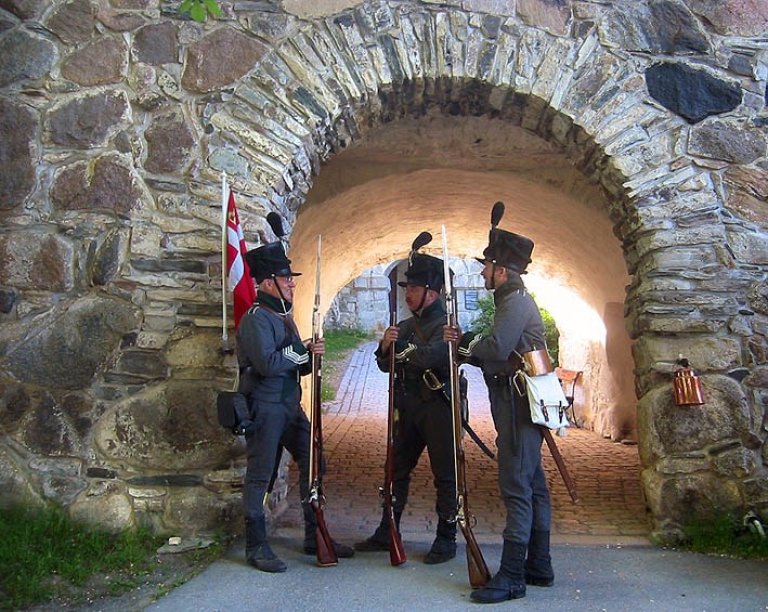 Tre personer i historiske militæruniformer under en steinbue, med rifler og et flagg, på en brosteinssti