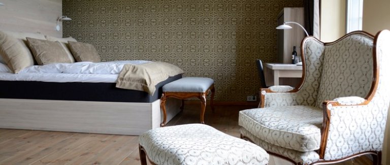 Velmøblert soverom med stor seng, lenestol, ottoman, og skrivebord med lampe, på lys tregulv