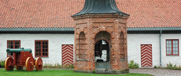 Historisk bygning med rødt tak og hvite vegger, med kanon på røde og gule hjul foran, og en brønnlignende struktur i midten