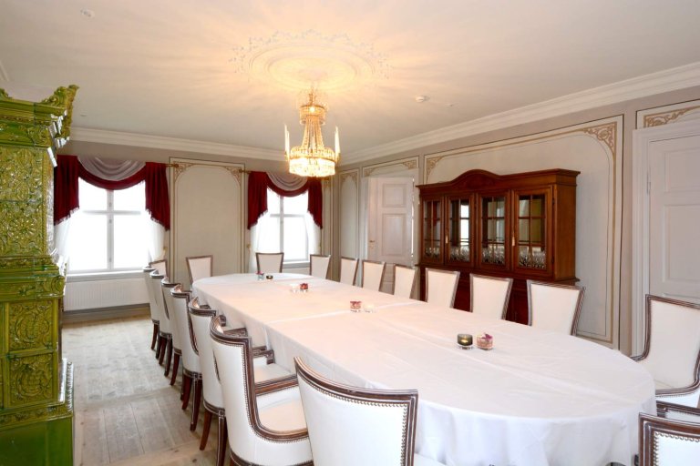 Elegant spisestue med lang ovalformet bord, hvite stoler med mørke treverk, lysekrone, store vinduer med røde og hvite gardiner, og grønn keramikkovn