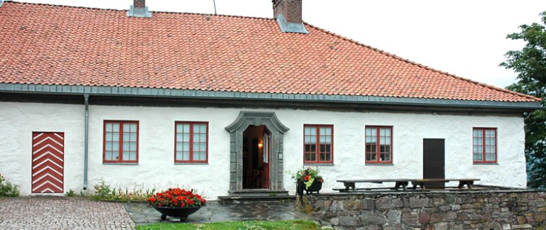 Hvitt hus med rødt tak og flere skorsteiner, med blomsterpotter og utemøbler på steinlagt terrasse