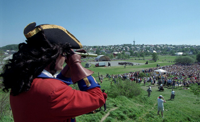 Person i historisk antrekk med tricorn hatt og rød frakk, som ser gjennom et teleskop på en stor folkemengde i et åpent felt