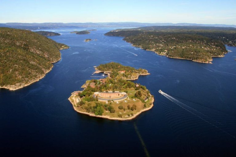 Luftfoto av kystlandskap med flere øyer, en stor rund struktur på den sentrale øya, og en båt som lager krusninger i vannet.