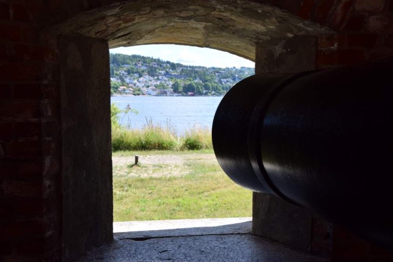 Utsikt fra innsiden av en steinstruktur med en stor svart kanon som peker mot et grønt område, vann og en åsside med hus og trær.