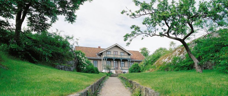 Malerisk hus med rødt tak, omgitt av frodig grøntområder og en steinvei som leder opp til huset.