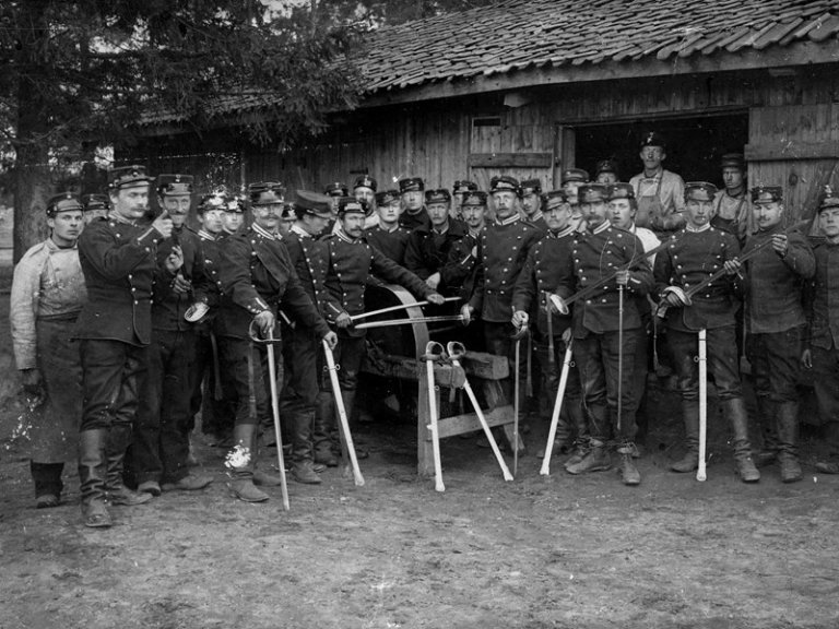 Historisk bilde av soldater i uniform med sverd foran en trebygning