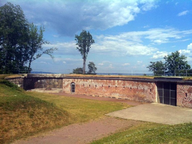 Utendørsscene med historisk murstruktur, med buet vegg, små rektangulære åpninger og buet døråpning, omgitt av gress og trær, med delvis skyet himmel.