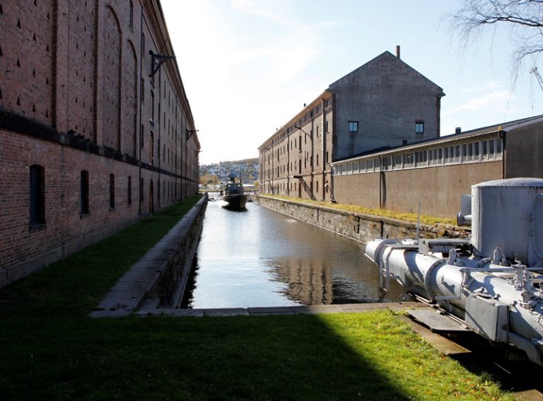 Smal kanal flankert av store murbygninger på begge sider, som leder til en vannkropp i det fjerne med en båt, omgitt av gress og utstyr i forgrunnen.