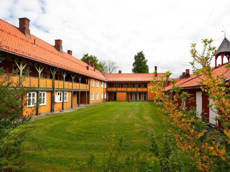 Gårdsplass omgitt av tradisjonelle trebygninger med oransje vegger og rødt tak, med balkonger på øverste etasje, velstelt plen og busker i bakgrunnen.