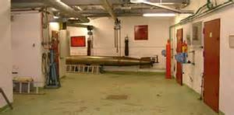 Industrielt rom med ulike maskiner og utstyr, inkludert rør i taket og en stor sylinder i midten.